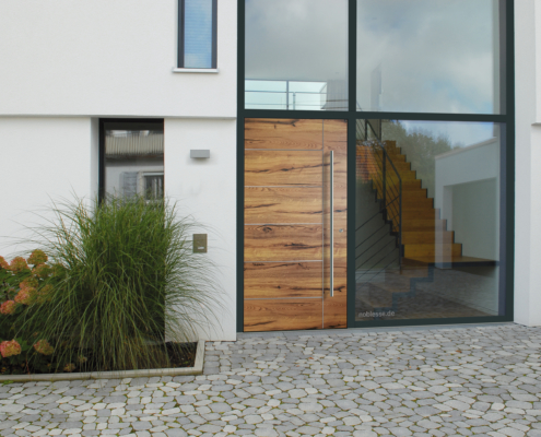 Ausschnitt eines Hauses, im Vordergrund Eingangstür aus Holz, links und darüber große Glaseinsetze